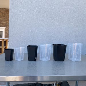Könnte beinhalten: Fünf Plastik-Blumentöpfe in verschiedenen Größen. Zwei sind schwarz und drei sind klar. Die Töpfe sind in einer Reihe auf einer silbernen Metalloberfläche angeordnet.