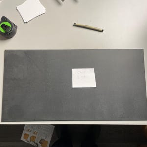 May include: A large, rectangular, dark gray surface with a textured appearance. A small white sticky note is attached, with handwritten text indicating dimensions of 12 x 24 inches and a thickness of 1/2 inch. A tape measure and pen are visible.