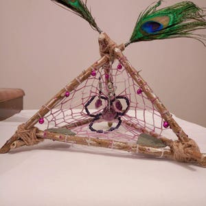 May include: A triangular dreamcatcher made of twigs and twine with pink and white netting. The dreamcatcher is decorated with purple beads, a purple crystal, and two peacock feathers.