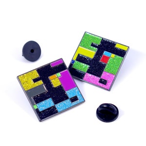 May include: Two colourful enamel pins with a geometric design. The pins are black with yellow, green, pink, blue, and red accents. The pins are glittery and have a square shape.