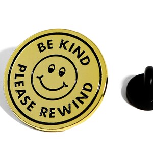 May include: Gold-colored metal pin with a black smiley face and the text "Be Kind Please Rewind".