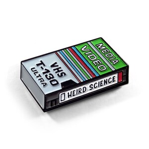 May include: A green, black, and white VHS tape enamel pin with the text "MEDIA VIDEO" and "WEIRD SCIENCE" on it.