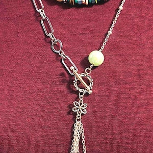 May include: A layered necklace with a beaded upper strand in green, yellow, and brown tones. The lower strand features a silver chain with a flower pendant and dangling charms, including a green bead and clear crystals.