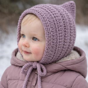 May include: A lavender-colored, crocheted baby bonnet with pointed ear details. The bonnet is tied with a matching lavender string. The baby is wearing a pink jacket with a hood. The background is a snowy, outdoor scene.