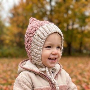 May include: A handmade crochet bonnet in soft pink and cream colors. The bonnet has a pointed top and ties under the chin. The child is wearing a beige jacket with a hood.