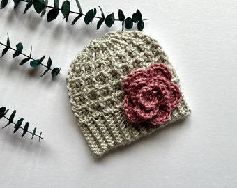 May include: A crocheted beanie hat in a light beige color with a pink crocheted flower on the side.