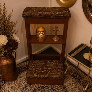 May include: A wooden prayer kneeler with a cushioned seat and shelf, featuring religious text. A gold cross and candle holder are on the shelf. A copper vase with flowers and a lit candle in a glass holder are also present.