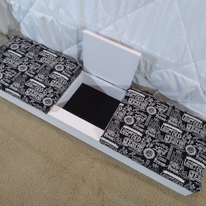 May include: A white bench with two cushioned seats and a storage compartment. The cushions are black with a white text pattern including the word "JESUS". The storage compartment is open, revealing a black interior.