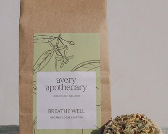Puede incluir: Una bolsa de papel marrón con una etiqueta blanca que dice "avery apothecary herb-infused wellness breathe well organic loose leaf tea". La bolsa está sentada sobre una tela azul claro con té de hojas sueltas esparcido a su alrededor. Hay un pequeño plato de cerámica con té de hojas sueltas y un pequeño adorno de cerámica de abeja.