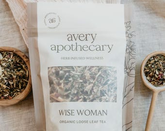 May include: A brown paper pouch with a white label that reads "avery apothecary HERB-INFUSED WELLNESS WISE WOMAN ORGANIC LOOSE LEAF TEA". The pouch is filled with loose leaf tea and is sitting on a light brown fabric surface.
