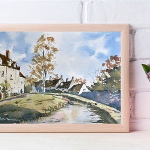 May include: Watercolor painting of a village scene in a light wood frame. The artwork depicts white buildings with brown roofs, a grassy area, and a body of water under a blue sky with clouds. A pink vase with purple flowers is in the foreground.