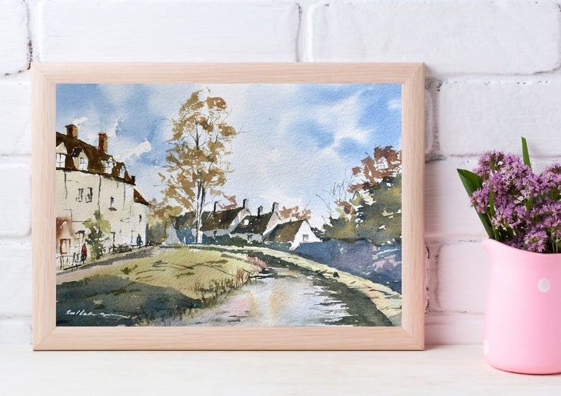 May include: Watercolor painting of a village scene in a light wood frame. The artwork depicts white buildings with brown roofs, a grassy area, and a body of water under a blue sky with clouds. A pink vase with purple flowers is in the foreground.