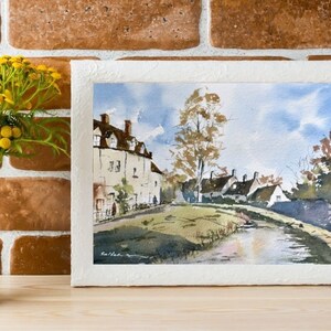 May include: Watercolor painting of a quaint village scene with white buildings, brown roofs, and a tree. The artwork is framed in a textured white frame. A white vase with yellow flowers sits to the left of the painting. The background is a brick wall.