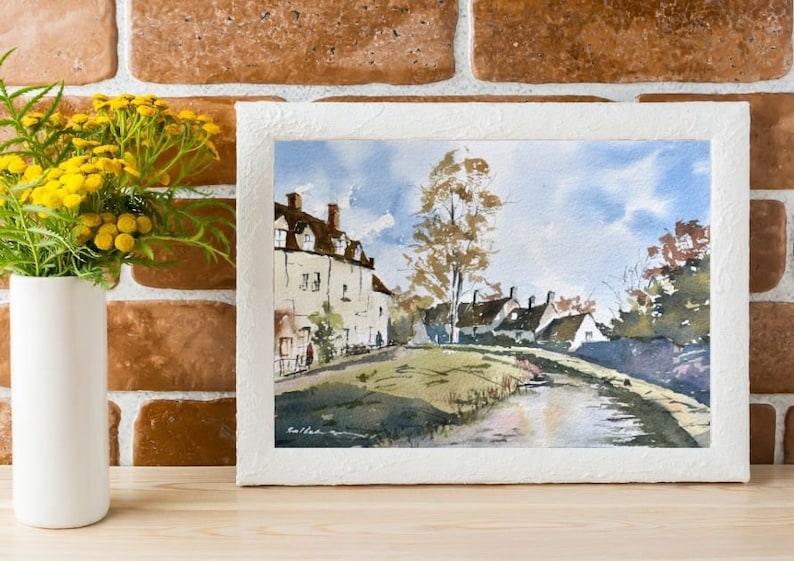 May include: Watercolor painting of a quaint village scene with white buildings, brown roofs, and a tree. The artwork is framed in a textured white frame. A white vase with yellow flowers sits to the left of the painting. The background is a brick wall.