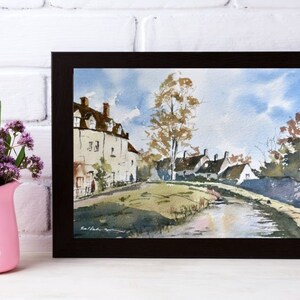May include: Watercolor painting of a village scene in a dark brown frame. The artwork depicts white buildings with brown roofs, a road, and a tree under a blue sky. A pink pitcher with purple flowers is on the left.