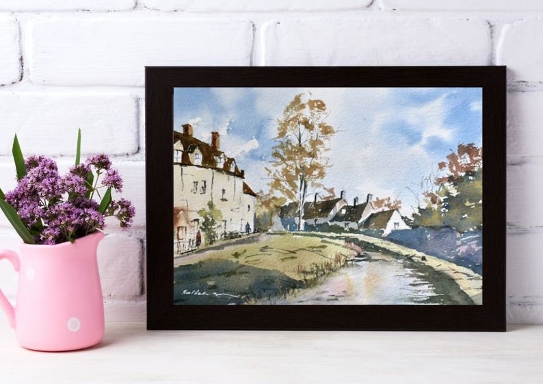 May include: Watercolor painting of a village scene in a dark brown frame. The artwork depicts white buildings with brown roofs, a road, and a tree under a blue sky. A pink pitcher with purple flowers is on the left.