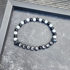 Può includere: Un braccialetto di perline bianche e nere con la scritta "ELDER EMO" in lettere bianche. Il braccialetto include perline a forma di stella ed è presentato su una superficie grigio scuro.