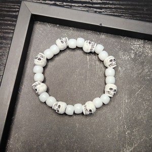 May include: White skull bead bracelet with a white background. The bracelet is made of white beads with a skull design.