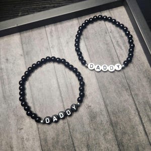 May include: Two black beaded bracelets, each spelling "DADDY" in white letter beads. The bracelets are made of small, round beads and are displayed on a wooden surface. A simple, elegant design, perfect for a gift.