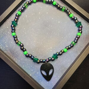 Puede incluir: Un collar de cuentas con un colgante de alienígena negro, cuentas iridiscentes, cuentas verdes en forma de estrella y cuentas verde brillante. El collar se muestra en una caja negra con un fondo estrellado.