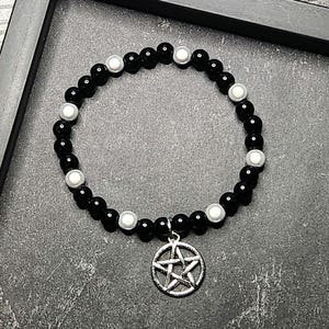 May include: A beaded bracelet featuring a repeating pattern of glossy black spherical beads and smaller white beads. A circular silver-toned metal charm with a five-pointed star pentagram design hangs from the bottom of the elasticized bead strand.