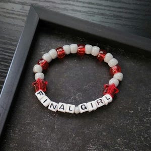 Könnte beinhalten: Ein kreisförmiges Perlenarmband mit weißen und transparenten roten Perlen, darunter zwei rote sternförmige Perlen. Das Armband enthält weiße quadratische Perlen mit schwarzer Beschriftung, die den Schriftzug FINAL GIRL bilden.