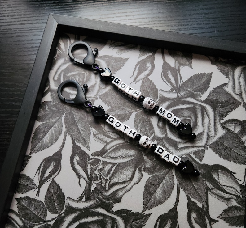 May include: Two black keychains with black and white beads spelling "goth mom" and "goth dad". The keychains are on a black and white floral background.