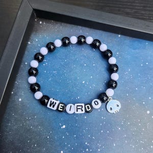 May include: A stretch bracelet featuring alternating black and white round beads. The bracelet includes white square letter beads spelling the word WEIRDO and a silver-toned metal charm shaped like a skull hanging from the beaded strand.