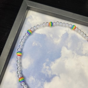 May include: A rainbow beaded necklace with iridescent clear beads and rainbow coloured beads. The necklace is on a white background with a blue and white cloudy pattern.