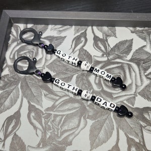 May include: Two black keychains with silver clasps. Each keychain has black and white letter beads spelling "GOTH MOM" and "GOTH DAD" with a skull bead between the words. The keychains are on a white and grey floral background.