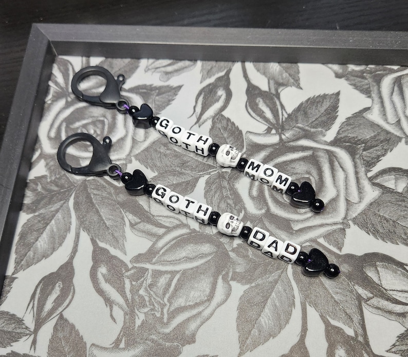May include: Two black keychains with silver clasps. Each keychain has black and white letter beads spelling "GOTH MOM" and "GOTH DAD" with a skull bead between the words. The keychains are on a white and grey floral background.