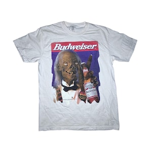 May include: White t-shirt featuring a graphic of a zombie holding a Budweiser beer bottle. The Budweiser logo is prominently displayed in red and white above the zombie image. The zombie is wearing a tuxedo. The shirt is a crew neck.