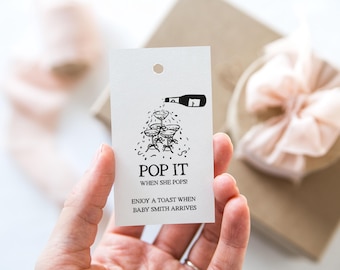 Può includere: Un'etichetta regalo bianca con testo nero e un disegno di bicchieri di champagne e una bottiglia. Il testo recita "Pop It When She Pops! Enjoy a toast when Baby Smith arrives."