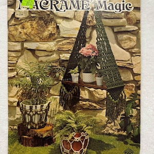 Könnte beinhalten: Ein Vintage-Buchcover mit dem Titel "Macrame Magic" mit einem Makramee-Pflanzenaufhänger mit Topfpflanzen. Das Cover zeigt einen Steinmauerhintergrund und eine Vielzahl von Pflanzen in Makramee-Haltern, die ein Retro-Bastelthema zeigen.
