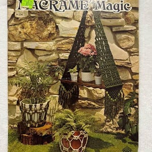 Può includere: Una copertina di un libretto di istruzioni Macrame Magic con un portapiante in macramè verde scuro con frange e perline, un portapiante in macramè nero e varie piante in vaso in supporti di macramè su una mensola e un ceppo.