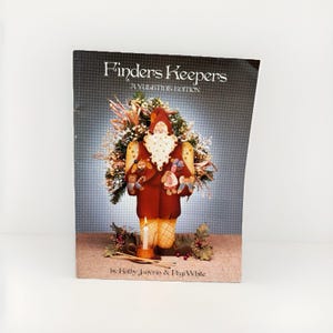May include: A softcover book titled Finders Keepers A Yuletide Edition by Kathy Janyrin and Pegi White. The cover features a brown-clad Santa figure with a white beard, surrounded by greenery, a lit candle, and small ornaments on a tan surface.