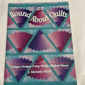 May include: A softcover book titled Round About Quilts by J. Michelle Watts. The cover features a quilt pattern with purple and pink fan-shaped designs on a light blue background. The text reads Unique Designs Using Wedge-Shaped Pieces.