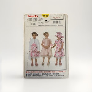 May include: A Burda 9648 sewing pattern envelope featuring illustrations of three outfits for children. The designs include a floral dress with a white bolero, a solid pink dress, and a floral dress with a matching sun hat. Text includes the brand name Burda, the pattern number 9648, and instructions in English, French, and Spanish.