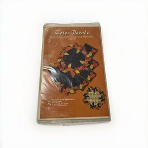 May include: A rectangular sewing pattern packet titled Color Parade Tablerunner, Table Topper and Placemats 050117 by Prairie's Edge Patchworks. The orange cover features illustrations of black fabric items decorated with colorful autumn leaf appliques.