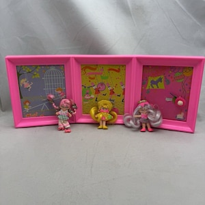 May include: A pink, three-panel picture frame with colorful illustrations and three small dolls. The frame has a bright pink finish. Each panel features a different scene with cartoon-like characters and designs. The dolls are positioned in front of the frame.
