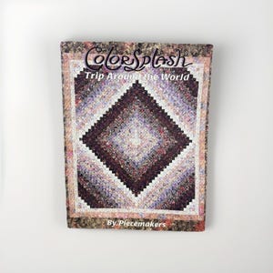 May include: A rectangular instructional book titled ColorSplash Trip Around the World by Piecemakers. The cover features a quilt with a diamond-shaped pattern in shades of purple, white, and dark brown, surrounded by a floral-patterned border.