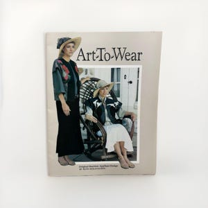 May include: A softcover instructional book titled Art-To-Wear by Ruth Seeley-Scheel featuring original machine-applique designs. The cover displays two photographs of individuals wearing hats and vests with applique details, set against a light tan background.