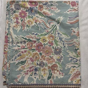 May include: A folded floral fabric featuring a bouquet of pink, yellow, and purple flowers on a light blue background. The fabric has a scalloped edge with a decorative trim.