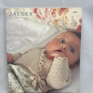 May include: A Jaeger Handknits pattern book, JB29, featuring a baby wearing a beige knit cardigan with a scalloped edge and tie closure. The cover includes the text: 27 designer garments by Martin Storey for babies & children using baby 4ply & baby DK.