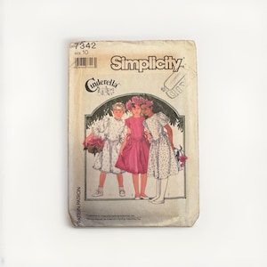 Op de afbeelding: Een Simplicity naaipatroon envelop, nummer 7342, maat 10, met een illustratie van drie figuren in jurken. De tekst bevat de merknaam Simplicity en het woord Cinderella. De envelop is gebroken wit met een gestippelde textuur.