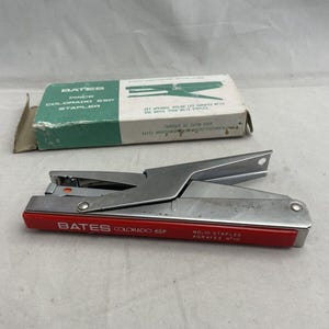 May include: A silver and red Bates Colorado 65P stapler with a matching box. The stapler has a red base with the text "BATES COLORADO 65P" and "NO. 10 STAPLES". The box is green and white.