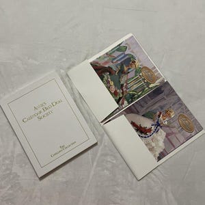 Op de afbeelding: Een wit boekje met de tekst "Annie's Calendar Bed Doll Society" en twee open brochures met poppenillustraties. De brochures zijn van "The Cotillion Collection."