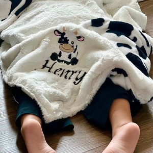 May include: A white and black cow print blanket with a white plush texture. The blanket has a small embroidered cow and the name "Henry" on it. A child's feet are visible under the blanket.