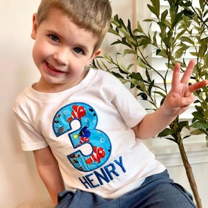 Op de afbeelding: Een wit T-shirt met een blauw nummer 3 geborduurd op de voorkant. Het nummer is omringd door kleurrijke vissen en bubbels. De naam "HENRY" is geborduurd onder het nummer.