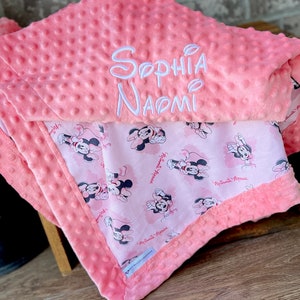 May include: A pink and white baby blanket with a Minnie Mouse print on one side and the name "Sophia Naomi" embroidered on the other side.